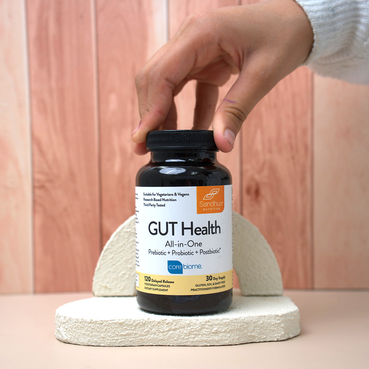 A hand holding a Sandhu Nutrition bottle labeled “Gut Health All-in-One” dietary supplement
