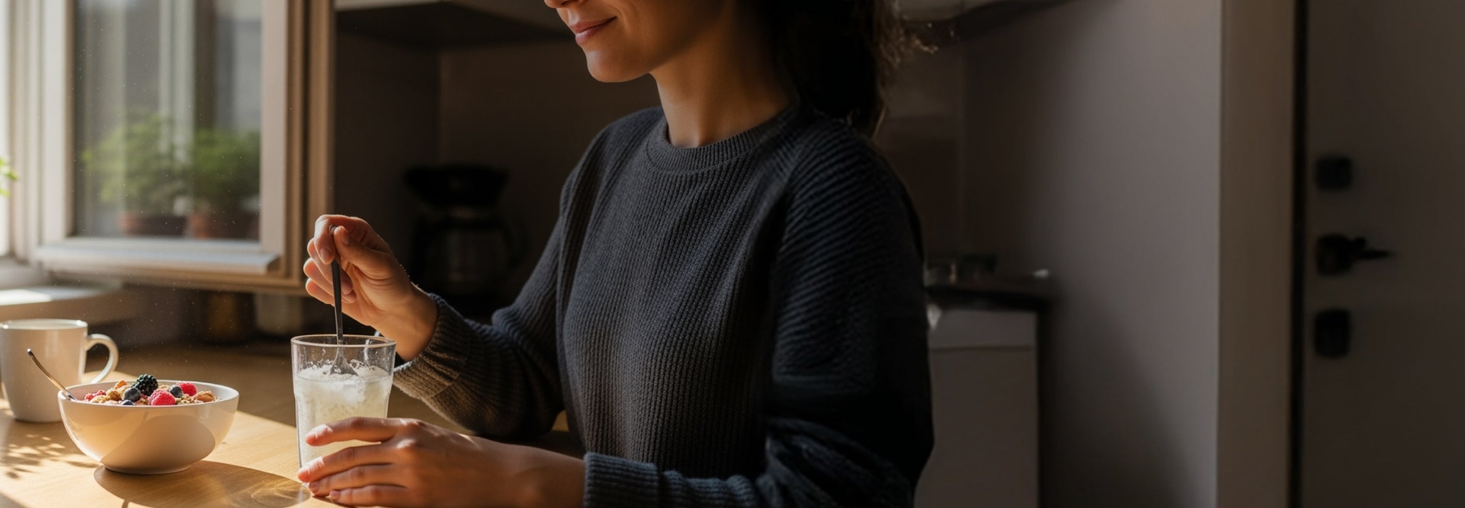 Person in a kitchen holding a glass of water with a bowl of fruit on a table.