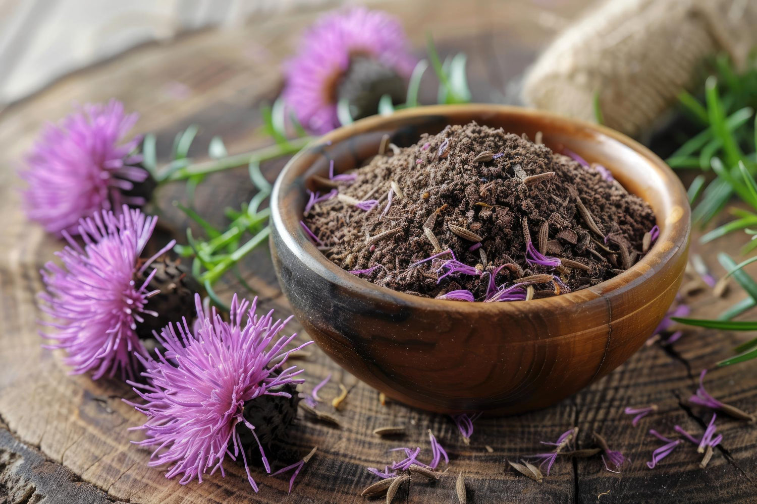 Wooden bowl filled with milk thistle powder surrounded by purple thistle flowers, a natural herb for liver health and detox support.