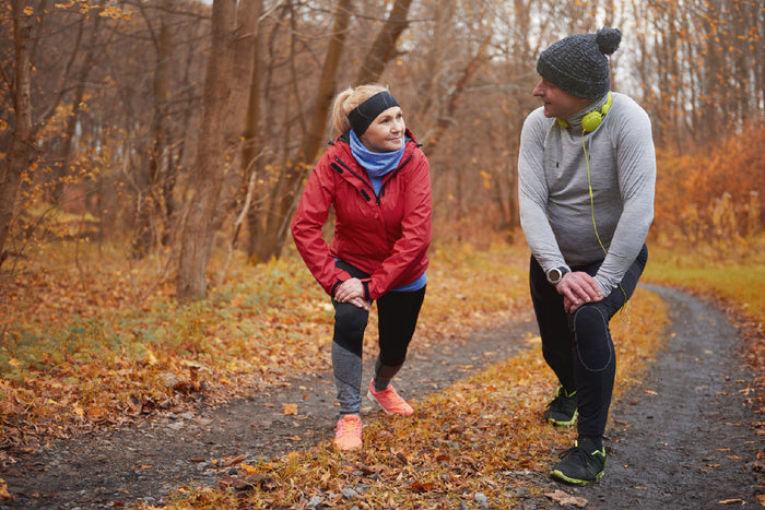 Active older couple stretching outdoors on a forest trail during fall, surrounded by colorful autumn leaves, preparing for a jog.
