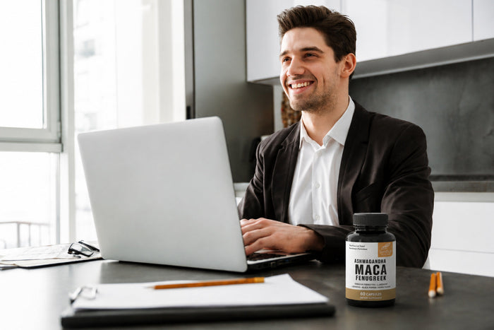 Smiling man works on a laptop beside Sandhus Ashwagandha Maca Fenugreek capsules, illustrating maca energy support for busy daily work.