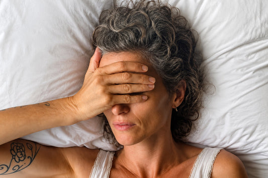 Middle-aged woman lying in bed with her hand covering her face, appearing tired and uncomfortable while resting on white bedding at night.