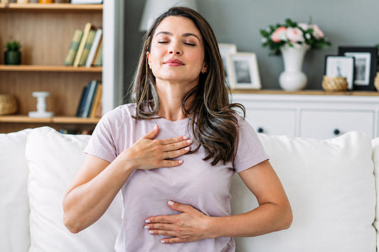 Woman sitting comfortably on a sofa with her eyes closed and hands resting on her chest and abdomen, demonstrating calm breathing and relaxation.
