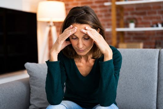 A middle-aged woman sitting on a sofa with her hands on her forehead, illustrating emotional stress, tension and changes in stress response during menopause.