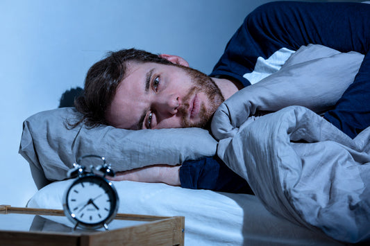 Man lying awake in bed with his eyes open and an alarm clock nearby, illustrating sleep difficulties caused by worrying about falling asleep.