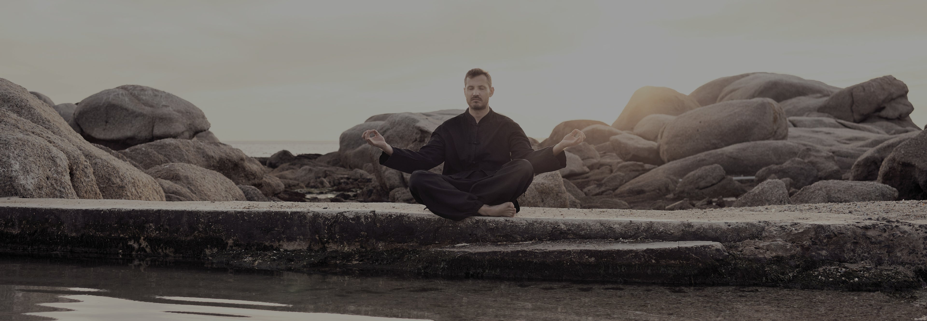 Person meditating on a rock by a body of water with large rocks in the background