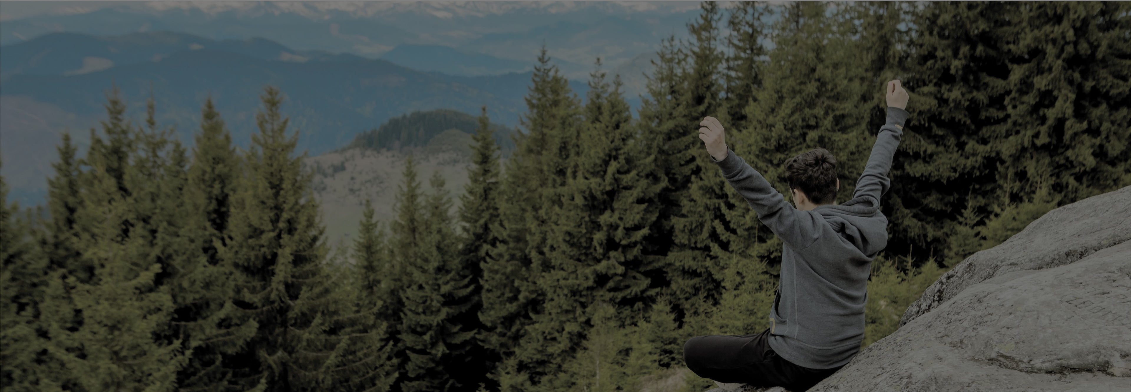 Person sitting on a rock with arms raised in a forest with mountains in the background